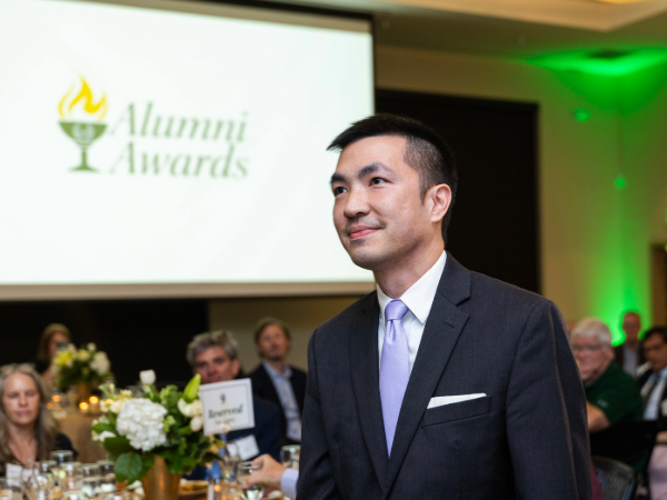 Man walking among others in an award ceremony setting with "Alumni Awards" on presentation in the background